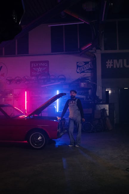 Mechanic working on a vintage car under neon lights in a dim garage setting.